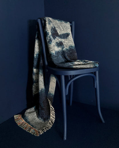 Draped over chair, silver gray and indigo blue woven cotton throw blanket with fringed edges. 54 by 72 inches. Against navy background.