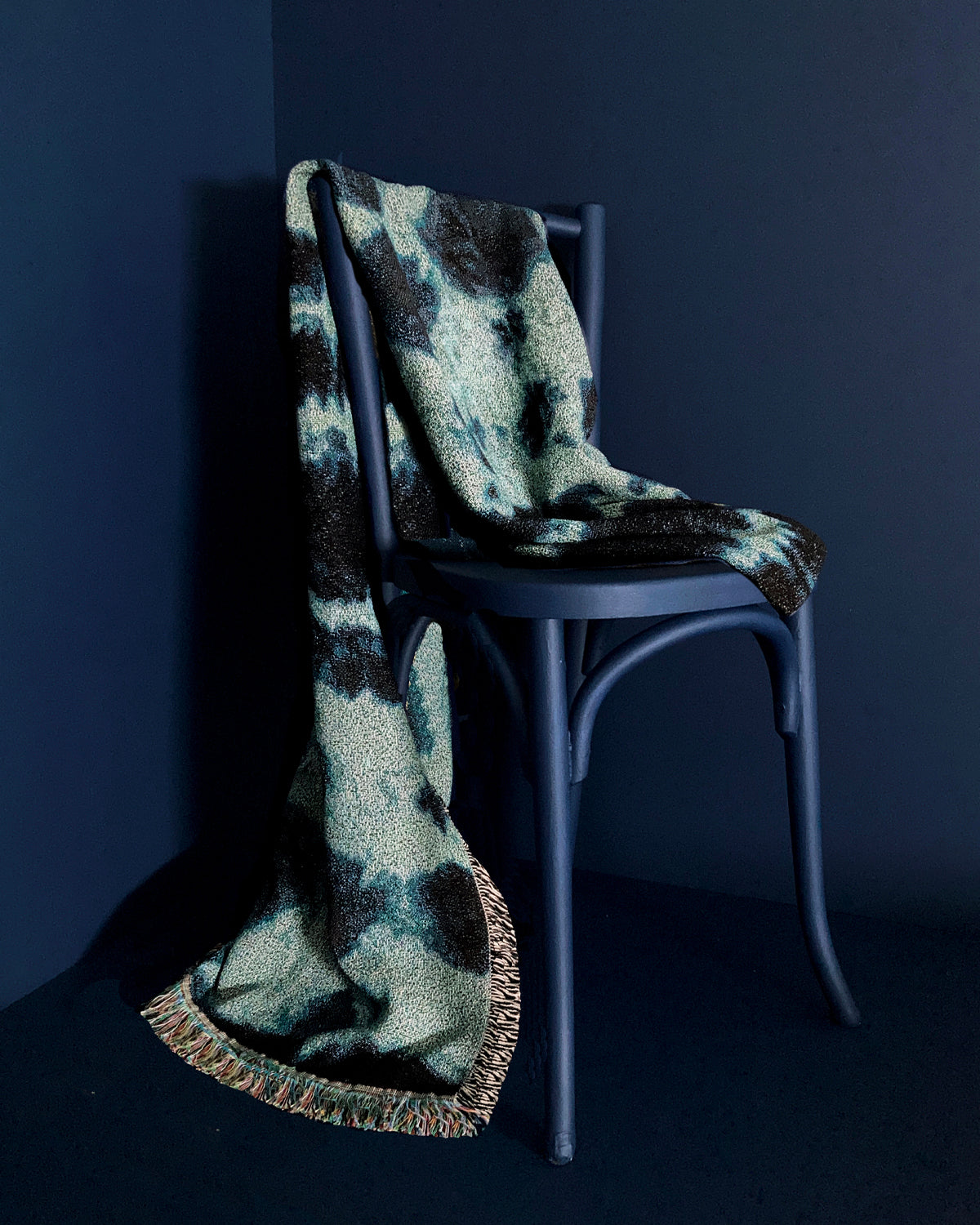 Draped over chair, jade green and indigo blue woven cotton throw blanket with fringed edges. 54 by 72 inches. Against navy background.